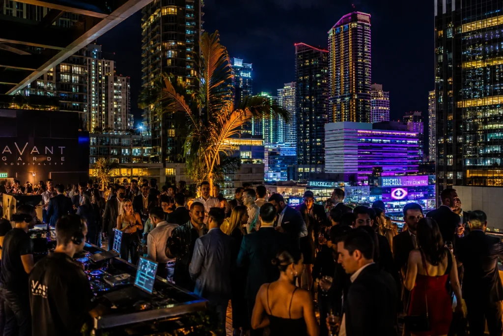 This image is showing a luxury Miami rooftop entertainment event with downtown skyline views, upscale nightlife atmosphere, and modern corporate celebration vibes.